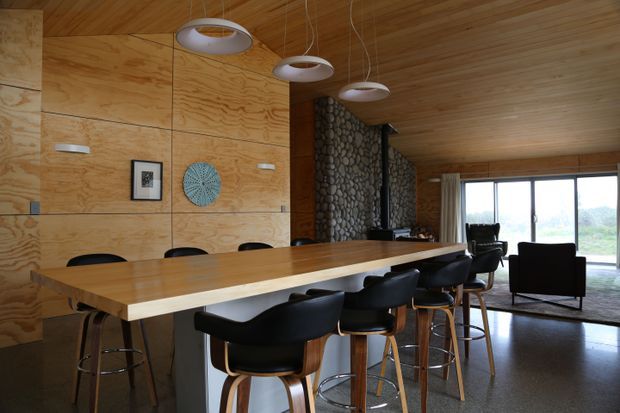 Open-plan dining and living area with a large timber dining table, black upholstered bar stools and chairs, plywood wall and ceiling lining, pendant lighting, a striking rounded-stone feature wall, and expansive glazing overlooking greenery. The space has a warm, contemporary natural-material palette.