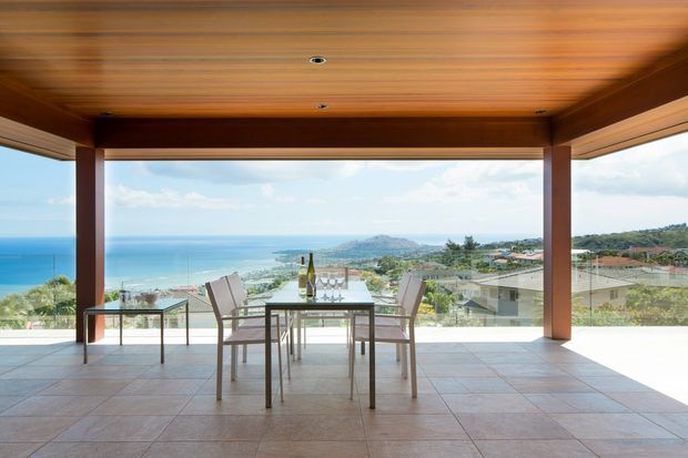 A covered outdoor terrace or balcony with a timber-lined ceiling, substantial timber posts, tiled flooring, and frameless glass balustrades. A dining table and chairs overlook a bright coastal bay and hillside settlement. The space is open, airy, and strongly connected to the landscape.