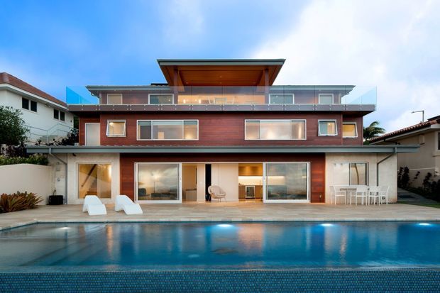 Contemporary multi-level residence viewed from the pool, featuring warm timber-clad walls, extensive glazing, broad terraces with glass balustrades, and a projecting flat roof over the upper level. The ground floor opens directly to the pool deck, with outdoor dining and lounge furniture visible.