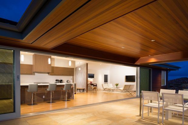 Evening view of a contemporary home with large open sliding glass doors connecting an outdoor entertaining area to a bright open-plan interior. The space features a dramatic timber-lined soffit and ceiling, warm timber flooring, a modern kitchen with island seating, and a living area beyond. Outdoor dining furniture occupies the foreground.