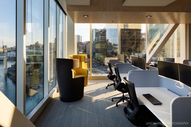 A bright contemporary office interior with floor-to-ceiling glazing overlooking a dense urban waterfront or city setting. The space contains rows of white and grey workstation desks with black office chairs, a small yellow lounge chair, carpeted flooring, exposed structural bracing, and a warm timber slatted ceiling with recessed lighting.