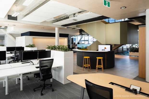 Bright contemporary open-plan commercial office interior with white workstations and black ergonomic chairs in the foreground, large built-in planters, a curved black reception or service counter with yellow stools, exposed ceiling services, timber wall panelling, and glazed circulation areas in the background.