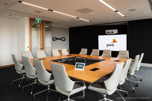 A contemporary corporate boardroom with a large rectangular conference table, numerous white swivel chairs, integrated power outlets, recessed linear ceiling lighting, and a wall-mounted presentation screen displaying the PwC logo. The room features a dark textured feature wall, glazed timber-framed entry doors, and carpet flooring.