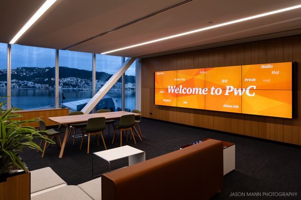 A contemporary corporate lounge and presentation space with floor-to-ceiling glazing overlooking a waterfront cityscape. The room features a large orange PwC digital display on a dark timber-panelled feature wall, integrated linear ceiling lighting, a meeting table with upholstered chairs, a sofa seating area, indoor plants, and dark carpet.
