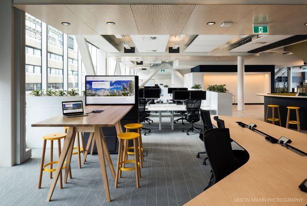 A bright contemporary open-plan office with floor-to-ceiling glazing, shared workstations, computer monitors, timber communal tables, stools, black office chairs, indoor plants, and a compact kitchenette or breakout bar. The ceiling combines timber slatted panels with acoustic and recessed lighting elements.