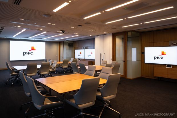 A modern corporate boardroom or conference room with a large central timber meeting table surrounded by upholstered swivel chairs. Multiple wall-mounted presentation screens display the PwC logo. The room features dark carpet, timber wall panelling and cabinetry, glazed partitions, acoustic ceiling panels, and linear recessed lighting.
