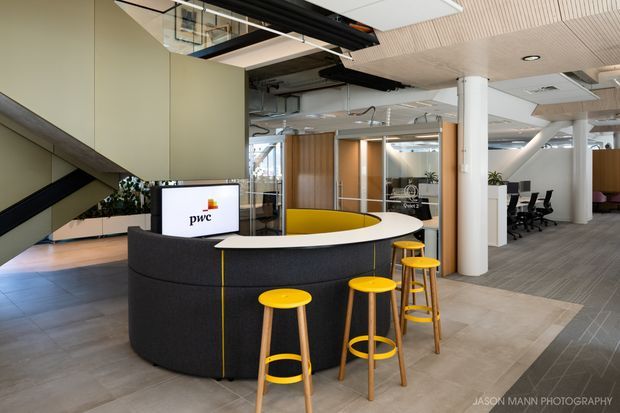 Modern open-plan commercial office reception area with a curved dark upholstered reception desk, yellow stools, a PwC-branded screen, exposed services, glass meeting rooms, white columns, and a mixed timber-slat and exposed ceiling. The space has a contemporary corporate fit-out with bold yellow accents.