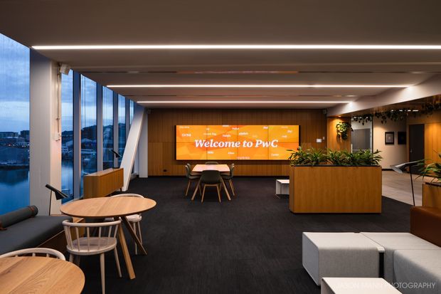 A contemporary commercial office reception or lounge area with floor-to-ceiling glazing, dark carpet, warm timber wall panelling and planters, integrated linear ceiling lighting, and flexible café-style seating. A large digital welcome screen identifies the space as a PwC workplace.