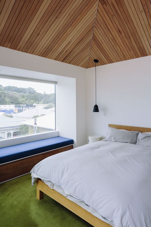 Bright contemporary bedroom with a large picture window, white walls, a warm timber-lined vaulted ceiling, and a simple timber bed. White bedding, a deep blue window seat, green carpet, dark timber built-in storage, and a black pendant light create a clean yet warm interior. A leafy suburban setting is visible outside.