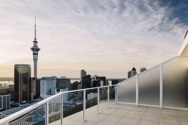 A rooftop terrace overlooking a dense urban skyline, with Auckland’s Sky Tower prominently visible in the distance. The terrace has pale tiled flooring, clear glass balustrades with white frames, and frosted privacy screens. The open city view and expansive sky create a bright, airy contemporary setting.