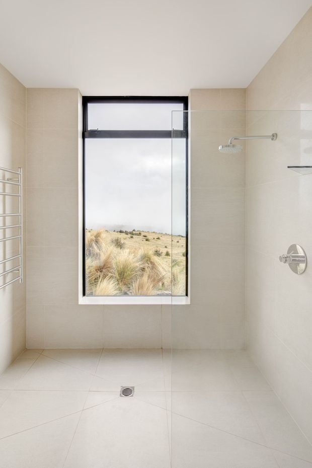 Bright, minimalist walk-in shower room with large pale beige tiled walls and floor, frameless glass shower screen, recessed chrome shower controls and showerhead, a partially visible heated towel rail, and a large black-framed window overlooking a dry grassland landscape. The room has a clean, contemporary fit-out with an open, airy feel.