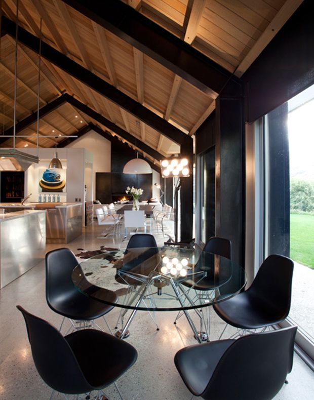 A dramatic open-plan dining and kitchen interior with a high pitched timber-lined ceiling, exposed dark structural beams, black-framed full-height glazing, and polished concrete flooring. A round glass dining table with sculptural black chairs occupies the foreground, while a contemporary kitchen, long dining area, pendant lighting, and fireplace extend into the background.