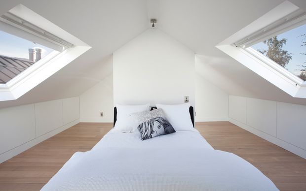 Bright, minimalist attic bedroom with a centered bed beneath a pitched ceiling. Large angled skylight windows on both sides bring in abundant natural light, while low built-in white cabinetry follows the roofline. The room has pale timber flooring, white walls and bedding, and a simple, uncluttered finish.