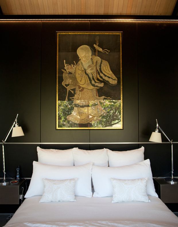 A refined bedroom with a large upholstered bed dressed in white and pale beige linens, set against a dark paneled feature wall. Matching angled bedside lamps sit on dark bedside tables, while a framed artwork is centered above the bed. A warm timber-lined ceiling and concealed linear lighting add warmth and contrast.