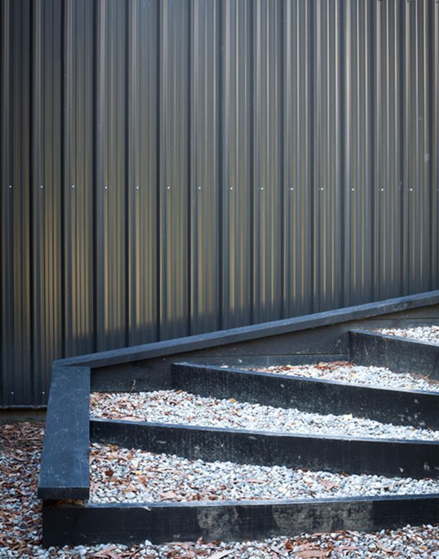 Close-up exterior view of a dark, vertically corrugated metal-clad wall beside broad outdoor steps. The steps and landing are finished in dark concrete or masonry and filled with loose pale gravel and leaf litter, creating a stark, linear contemporary entrance composition.