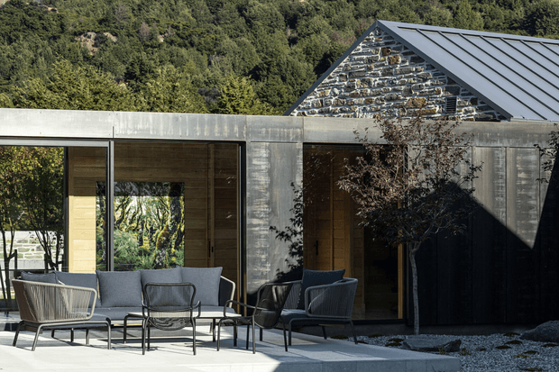 Contemporary single-storey residence with a low, rectilinear concrete-clad extension connected to a steep gabled structure featuring exposed dark stone masonry and a standing-seam metal roof. Full-height glazing opens onto a paved outdoor seating area furnished with modern lounge chairs. Dense wooded hillside surrounds the property.