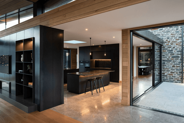 A sophisticated open-plan kitchen and living area with a large black island, integrated dark cabinetry, warm timber ceiling and wall framing, and polished stone flooring. Pendant lights illuminate the island, while full-height glazing opens to a paved courtyard and reveals a stone-clad exterior wall and adjoining dining area.