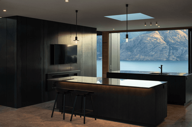 A dark, contemporary kitchen with a large island and integrated seating, black cabinetry, a reflective dark benchtop, and pendant lighting. Full-height glazing opens to a dramatic mountain and water view, while a skylight brings additional daylight into the space.
