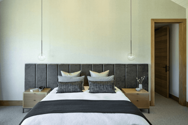A refined bedroom with a centered bed dressed in white linens and layered charcoal-grey cushions and throw, set against a full-height dark upholstered panelled headboard. Matching timber bedside tables flank the bed, with simple pendant lights suspended on either side. A timber-framed doorway with a horizontal timber-panel door opens to an adjoining space. The room has pale walls, neutral carpet, and a restrained contemporary palette.