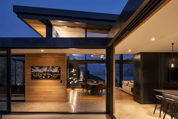 A dramatic dusk view into a contemporary open-plan home through expansive floor-to-ceiling glazing. Warm timber wall panelling and polished concrete floors frame a dining area and living room with mountain views, while a dark sculptural kitchen island and a covered rooftop or upper-level terrace create strong horizontal architectural lines. Integrated lighting and timber soffits add warmth against the dark exterior structure.