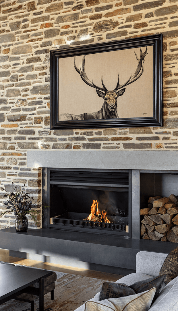 A cozy living room focused on a substantial open fireplace set into a textured natural-stone wall. The fireplace has a broad grey stone mantel and dark built-in hearth cabinetry, with stacked firewood in an adjacent niche. A framed deer artwork hangs above the mantel, while a contemporary sofa, coffee table, rug, and dried botanical arrangement occupy the foreground.