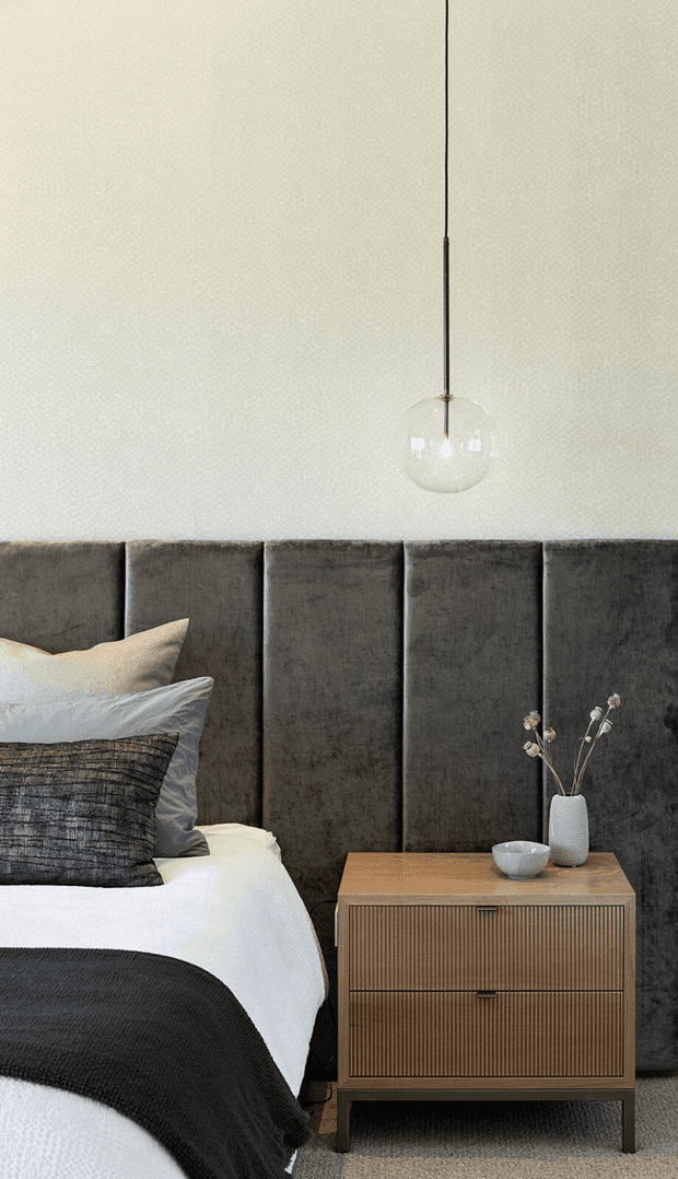 A close-up view of a refined bedroom featuring a tall charcoal upholstered panelled headboard, layered white and charcoal bedding, and a warm timber bedside table with fluted drawer fronts. A simple globe pendant hangs against a subtly textured pale wall, with understated ceramic accessories and dried stems adding a calm, contemporary feel.