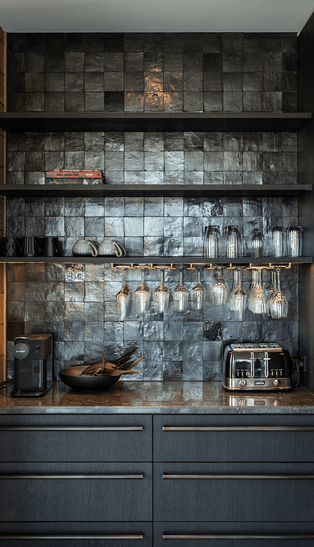 A close-up view of a dark, refined kitchen or bar joinery wall featuring open shelves lined with handmade textured dark tiles. Hanging stemware, tumblers, ceramic vessels, a coffee machine, toaster, and bowl sit above dark vertically grained cabinetry with long metallic drawer pulls. The palette is moody charcoal, black, slate grey, and warm bronze, with reflective highlights creating a luxurious atmosphere.