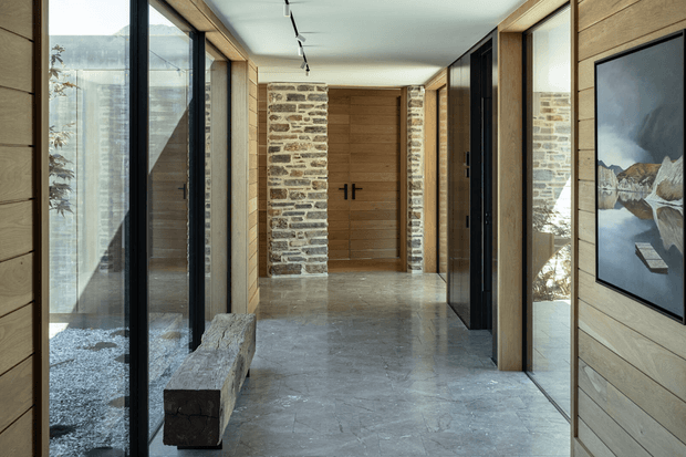 A refined modern hallway or entry corridor with full-height glazing on one side, pale timber wall panelling, exposed stone masonry, dark-framed internal doors, and a grey natural-stone floor. A built-in stone bench sits beside the glazing, while a large framed landscape artwork adds visual interest. The corridor has a calm, minimalist alpine-inspired character with strong natural materials and clean linear detailing.