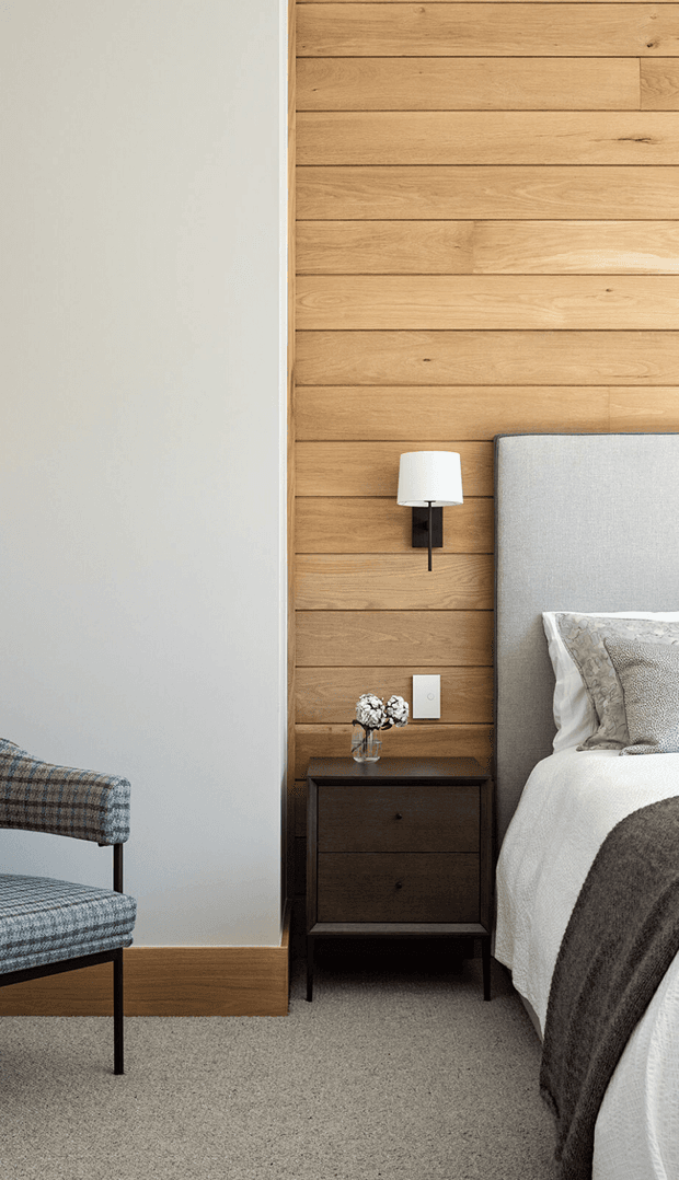 A refined bedroom corner with a warm horizontal timber feature wall, upholstered grey headboard, white bedding, dark wood bedside table, wall-mounted sconce, and a patterned accent chair. Neutral carpet and pale plaster walls create a calm, modern interior with restrained natural materials.
