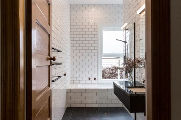 A bright, narrow bathroom viewed through an open timber door. White rectangular subway tiles cover the walls and bathtub surround, with dark grey floor tiles, a freestanding-style bathtub beneath a window, and a dark floating vanity with a stone benchtop and wall-mounted fixtures. A frameless glass shower screen and black tapware create a clean, contemporary look.