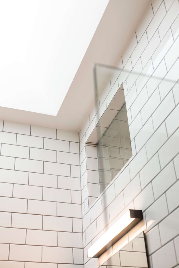Close-up view of a bright, contemporary bathroom corner clad in white rectangular subway tiles with dark grout. A large frameless mirrored cabinet or mirror is mounted on the tiled wall, with a slim linear wall light beneath it. The ceiling has a broad recessed or angled white plane, creating an airy, clean-lined composition.