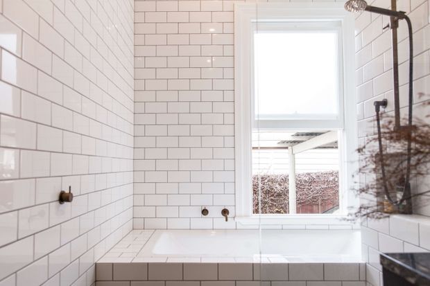 Bright, compact bathroom with a white tiled bathtub surround and white subway-tiled walls. A white-framed window provides natural light, while dark bronze or black shower hardware and taps add contrast. The room has a clean, simple contemporary-traditional appearance.
