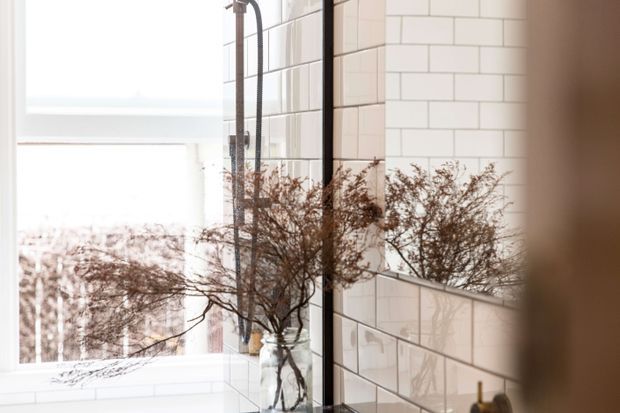 Bright contemporary bathroom with glossy white rectangular subway-tile walls, a frameless glass shower enclosure, and a large mirror reflecting delicate dried-branch arrangements. The space has a clean, light-filled appearance with dark shower hardware and subtle warm accents.