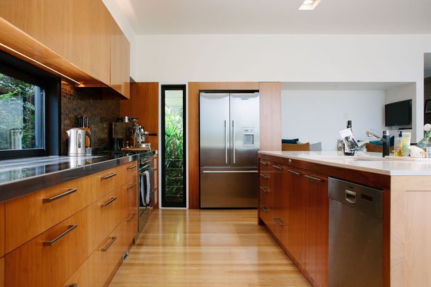 A bright, spacious contemporary kitchen with warm timber cabinetry, extensive dark stone benchtops, stainless-steel appliances, and a large white island or peninsula. The room includes abundant built-in storage, timber flooring, a refrigerator wall, dishwasher, sink, and views to greenery through the window and glazed door.