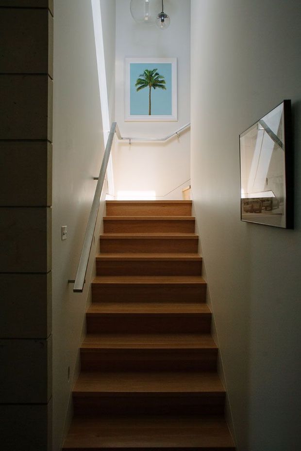 A narrow interior stairway with warm timber treads and risers, white painted walls, and simple slim metal handrails. The stair rises toward a bright landing with a framed palm-tree artwork and a small pendant light. A large framed mirror or reflective artwork is mounted on the right wall, while dark built-in joinery is visible along the left edge.
