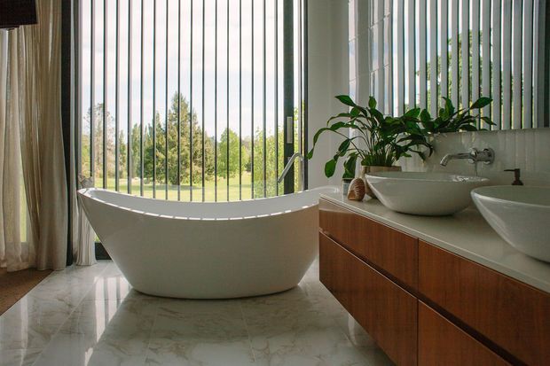 A bright, spacious bathroom with a freestanding white bathtub positioned beside full-height windows with vertical blinds or screening. A double vanity with white vessel basins, chrome fixtures, and warm timber cabinetry runs along the right wall. Large pale marble-look floor tiles and indoor plants contribute to the refined, natural atmosphere, with leafy grounds visible beyond the glazing.