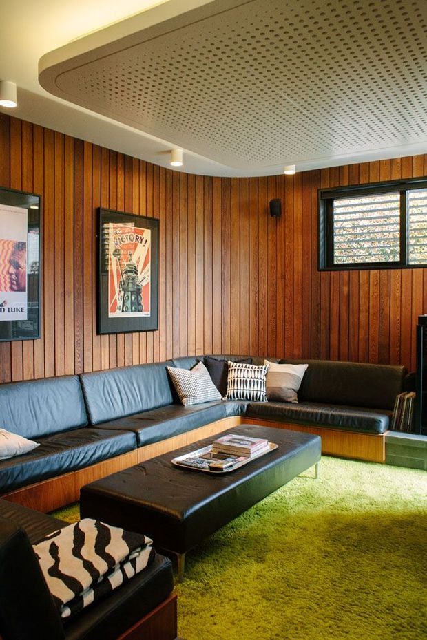A retro-inspired living room with extensive vertical timber wall panelling, a low black leather sectional sofa, a large black upholstered ottoman coffee table, patterned cushions, framed artwork, and a vivid green shag rug. The room features a perforated acoustic ceiling panel with rounded detailing, recessed spotlights, and dark-framed horizontal windows.