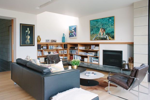 A bright, mid-century-inspired living room with a large black leather sofa, a leather-and-metal lounge chair, a round low coffee table, timber flooring, and extensive built-in timber shelving. A long timber mantel and white fireplace surround form the main feature wall, with artwork and books adding colour and character. The room opens toward a darker tiled hallway and is illuminated by a simple linear ceiling light.