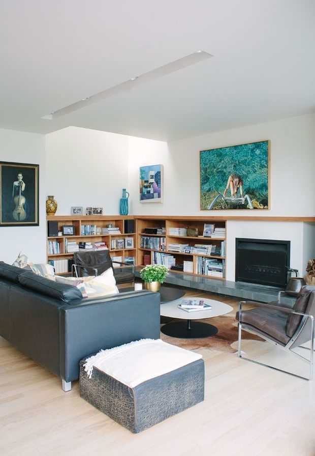 Bright, well-appointed living room with pale timber flooring, white walls and ceiling, a long built-in timber bookcase and display shelving, and a dark stone or concrete fireplace hearth with a black recessed fireplace. The room contains a large dark leather sofa, matching ottoman, leather lounge chairs, and a low round coffee table. Framed artwork and books add visual interest. A slim linear ceiling light reinforces the clean, understated mid-century-influenced interior.