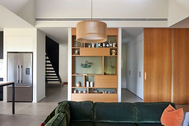 Contemporary open-plan living area viewed toward a built-in timber display and storage wall. A deep green sectional sofa occupies the foreground, with a large warm wood pendant light above. The space includes a stainless-steel refrigerator and kitchen area at left, a dark open stairway, pale tiled flooring, white walls and ceiling, and full-height timber cabinetry or sliding doors at right.