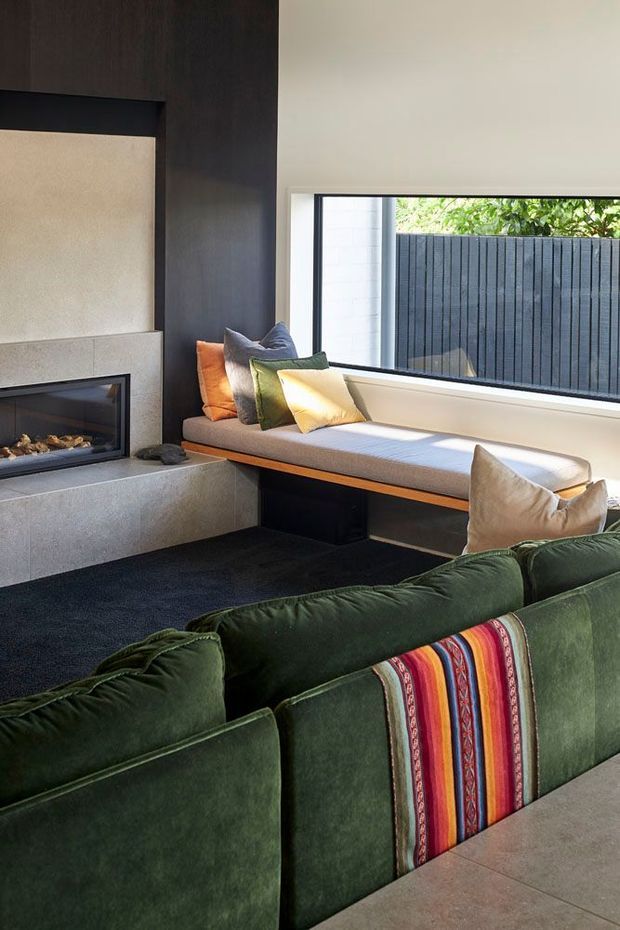 A refined living room with a built-in linear fireplace set into a dark timber feature wall, a long upholstered window seat, and a deep green sectional sofa with a colourful striped textile accent. Large horizontal glazing overlooks a dark vertical privacy fence, while dark carpet and pale stone or concrete surfaces create a strong contrast. The room is warmly lit and carefully furnished with earthy green, rust, mustard, and neutral tones.