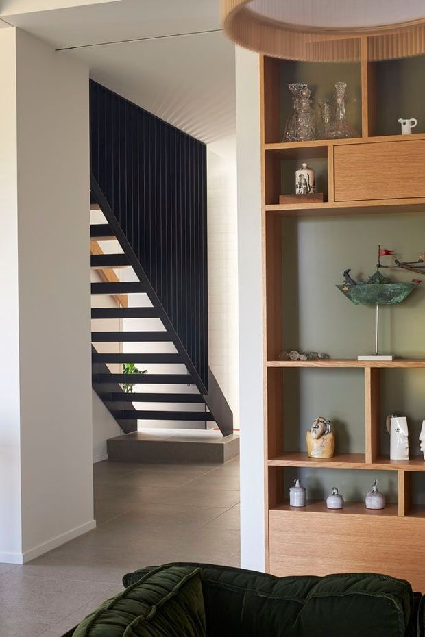 Interior view of a refined contemporary living area and stair hall. A floating open-riser staircase with a dark vertical slatted balustrade rises beside pale walls and large-format neutral flooring. A tall built-in timber display cabinet with open shelving and drawers occupies the right side, showcasing decorative objects. The foreground includes part of a deep green upholstered sofa, while a large layered pendant light is visible overhead. The space has a warm, curated and understated character.