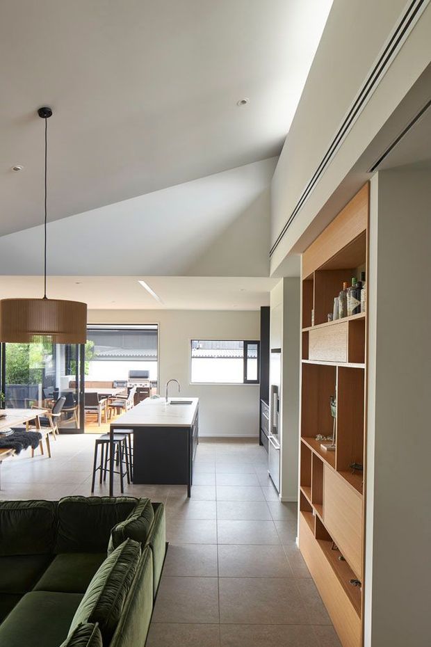 A bright open-plan interior showing a contemporary kitchen, dining area and living space. The kitchen has a long island with a pale benchtop, dark cabinetry and stools, while a full-height timber display and storage unit lines the right wall. A green upholstered sofa occupies the foreground, and large windows and glazing connect the dining area to an outdoor entertaining space. The room features a high angled ceiling, recessed lighting and a large pendant over the dining table.