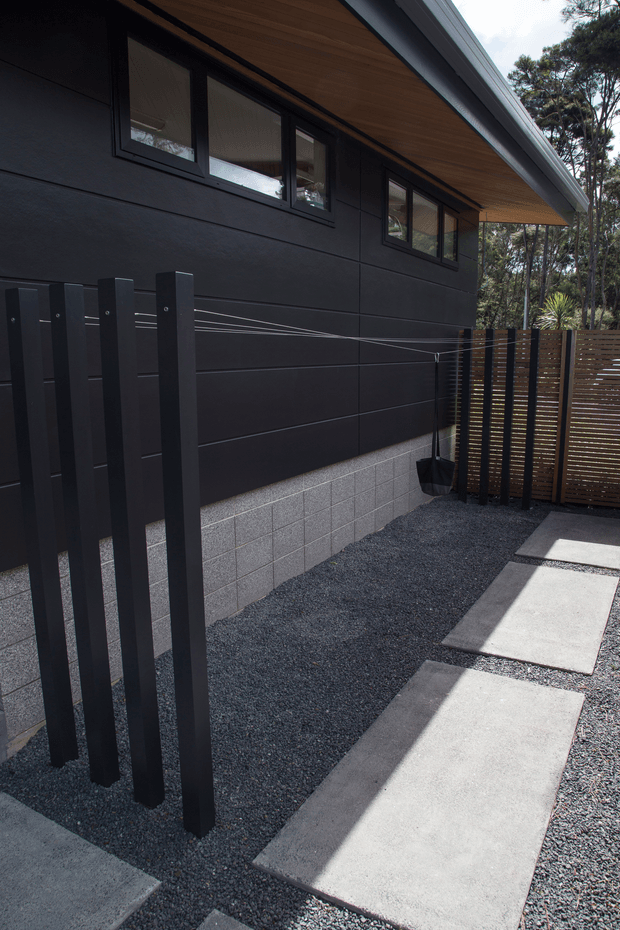 A contemporary residential exterior side passage with dark horizontal cladding, black-framed windows, and a warm timber-lined soffit beneath a broad roof overhang. The ground is finished with dark gravel and large rectangular concrete stepping slabs, bordered by a slatted timber privacy fence and vertical dark posts. Dense trees are visible beyond the boundary.
