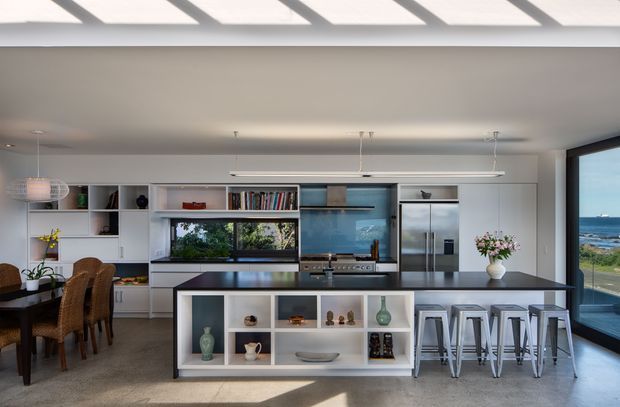 Bright, open-plan contemporary kitchen with a large central island, integrated stainless-steel appliances, white built-in cabinetry, open display shelving, and bar seating. A dining area sits to the left, while full-height glazing on the right overlooks a coastal ocean view. The room has a clean, airy feel with strong natural light and a neutral white, grey, and dark benchtop palette.