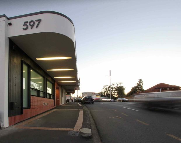 Street-level exterior view of a contemporary commercial building at 597, featuring a curved white projecting canopy, dark vertical cladding, large glazed windows, exposed red brick at the lower facade, and recessed linear lighting over the footpath. The building fronts a paved urban roadway with neighboring buildings visible across the street.