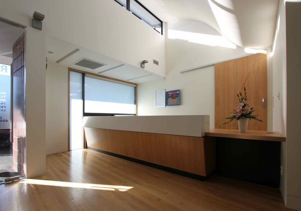 Bright contemporary reception or entry area with a built-in timber reception desk, white counter, dark base, timber feature wall, polished timber flooring, large glazing, and a high ceiling with clerestory-style windows and angular skylight-like openings. A floral arrangement and framed notices sit on the fixed reception joinery.