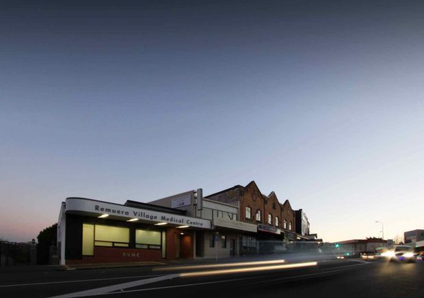 Dusk exterior photograph of the Remuera Village Medical Centre, a low-rise commercial building on a street corner. The foreground shows a broad paved roadway with light trails from passing vehicles, while the building features a curved projecting entrance canopy, illuminated glazing, dark brick walls, and an adjoining row of older-looking gabled brick facades.
