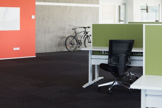 A contemporary open-plan commercial office interior with white workstations, green privacy screens, black ergonomic office chairs, and dark carpet flooring. A vivid coral-red feature wall and exposed concrete wall add colour and texture, while bicycles are stored against the rear wall.