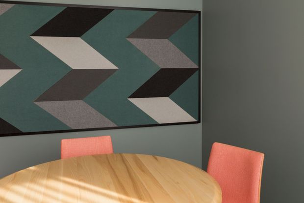 A dining area with a round light timber dining table and coral upholstered dining chairs, set against dark grey-green walls. A large framed geometric artwork with muted teal, charcoal, beige and grey shapes dominates the wall, creating a mid-century-inspired focal point. Strong sunlight casts linear shadows across the table.